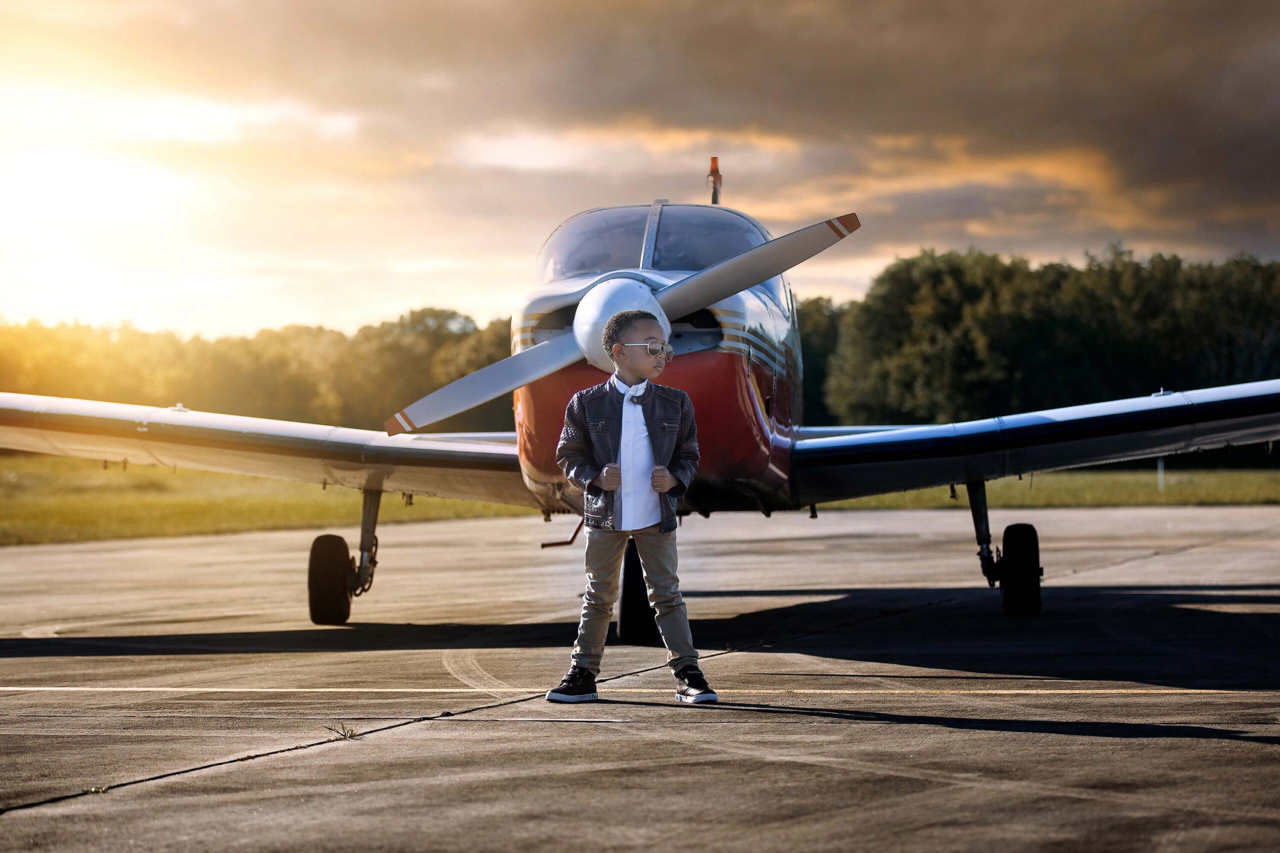 Adorable boy dressed in a leather ujacket wearing aviator sunglasses posing in front of an airplane